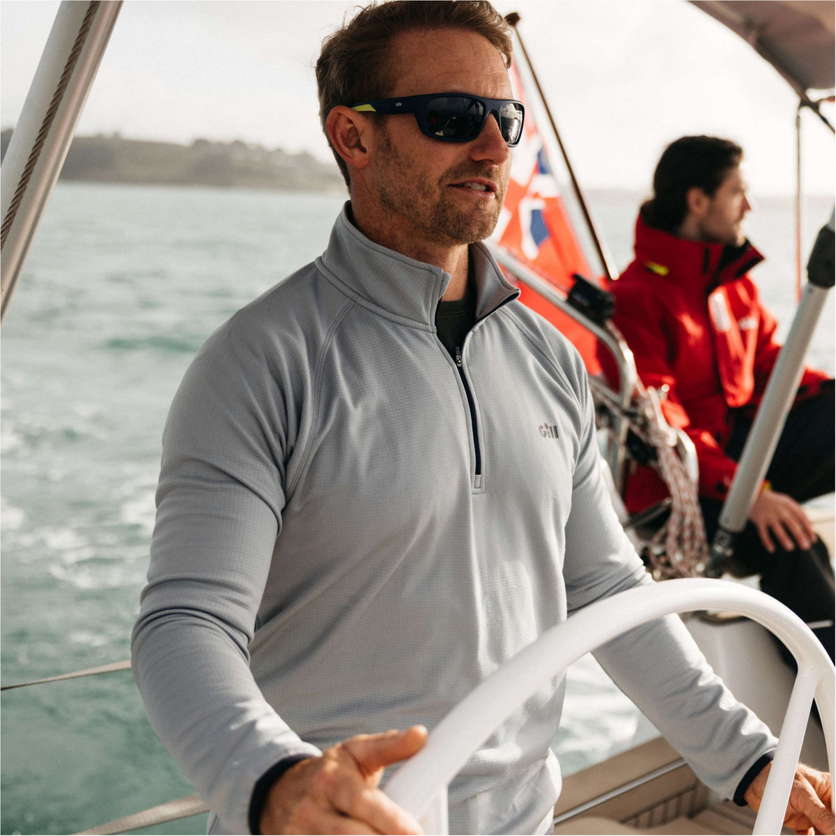 A man wearing a light gray long-sleeve quarter-zip performance top and black sunglasses is steering a sailboat on the water, with another person in a red jacket visible in the background.