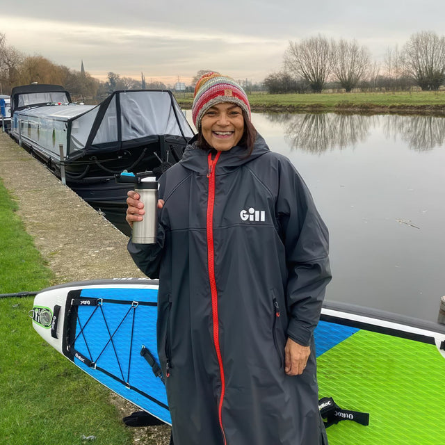 Person in a Gill raincoat holding a mug by a canal with boats in the background