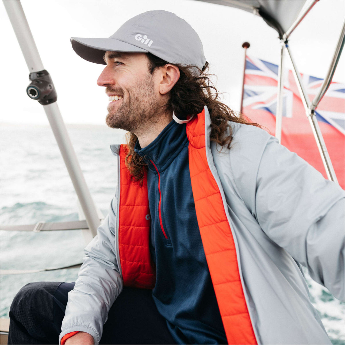 A man wearing a light gray GHI brand cap, a red and navy quilted vest over a navy shirt, and a light gray jacket, sitting on a sailboat with the ocean and a Union Jack flag in the background.
