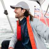 A man wearing a light gray GHI brand cap, a red and navy quilted vest over a navy shirt, and a light gray jacket, sitting on a sailboat with the ocean and a Union Jack flag in the background.