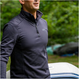 Man wearing a dark gray jacket with a brand logo, standing outdoors with greenery in the background.
