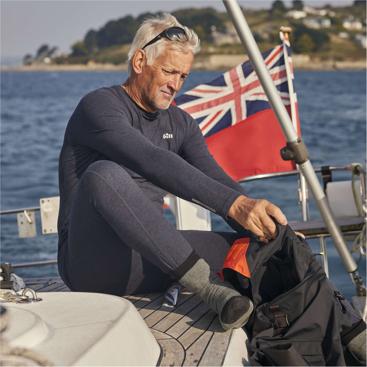 A man wearing a dark gray full-body wetsuit sits on a boat deck by the water, packing a black bag. A Union Jack flag is visible in the background.