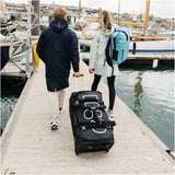 Two people walking away on a dock by the water, with one pulling a large black rolling duffel bag and the other carrying a turquoise backpack. The dock is surrounded by boats and water, with buildings visible in the background.