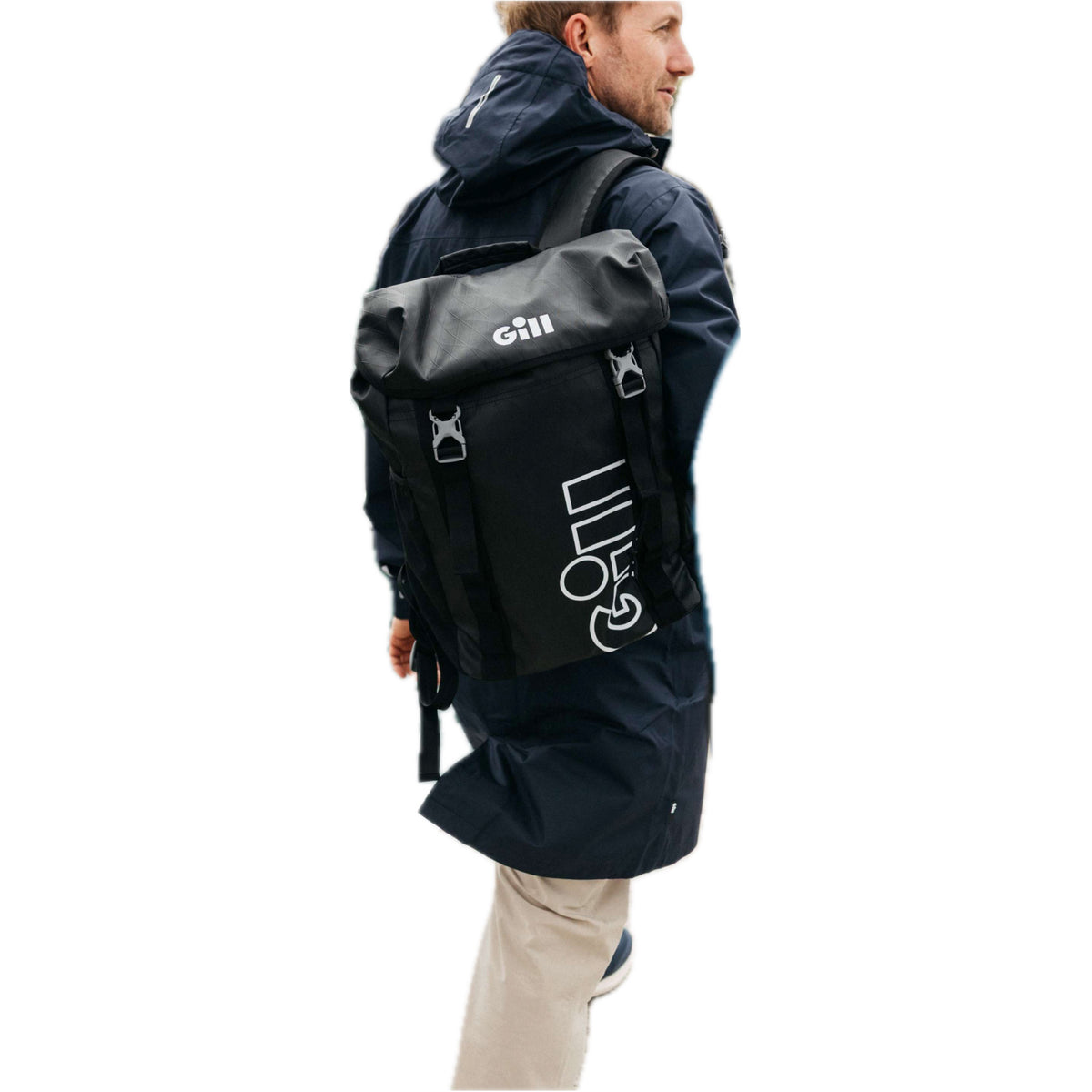 A man wearing a dark navy hooded jacket and beige pants, shown from the side/back, carrying a black Gill pack with white branding on a plain white background.