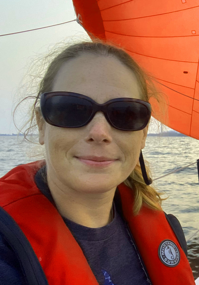 Person wearing sunglasses and a red life jacket on a boat with an orange sail.